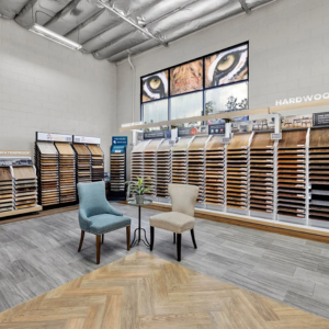 Flooring showroom displaying various hardwood and waterproof flooring samples, with two chairs placed in the center and a large tiger eye mural above the display.